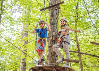 Mother and son climbing in extreme road trolley zipline in forest on carabiner safety link on tree to tree top rope adventure park. Family weekend children kids activities concept.